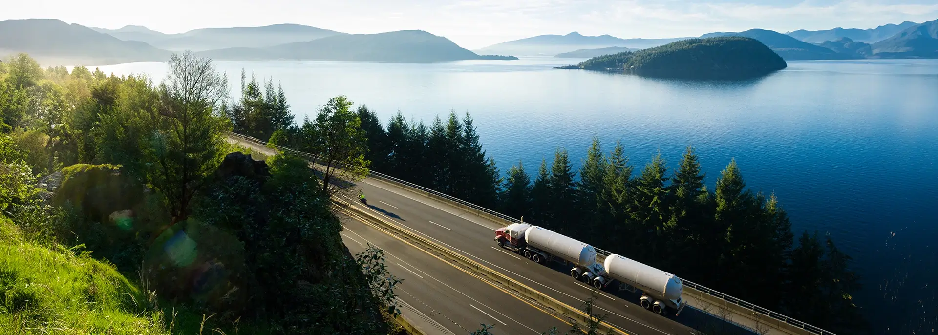 Downward view of tanker truck driving down a road near pine trees and water