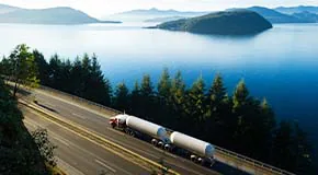Downward view of tanker truck driving down a road near pine trees and water