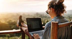 Person sitting on a balcony with feet up and typing on a laptop