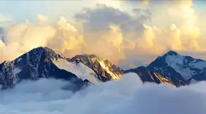 Wide hero shot of glowing, snow-covered mountains peaking over a blanket of clouds during sunrise
